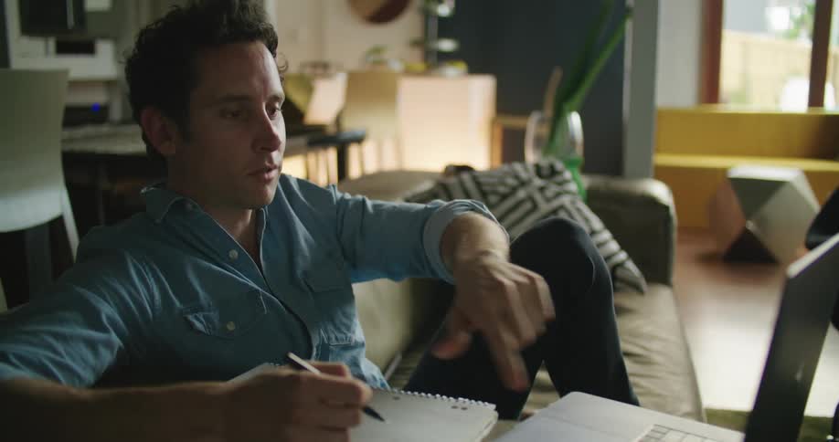 A man in a room sitting on a couch with a pen and a book and video chatting on a laptop