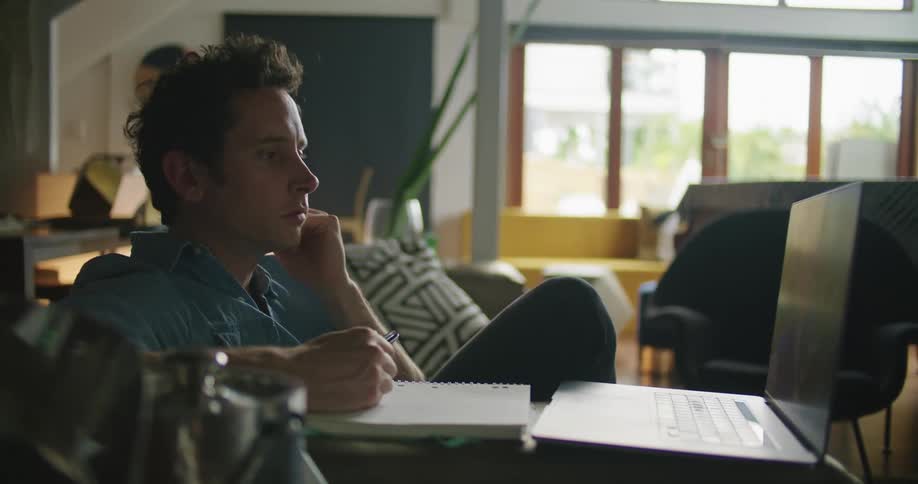 A man taps his notebook and gestures as he talks during a video call on his laptop