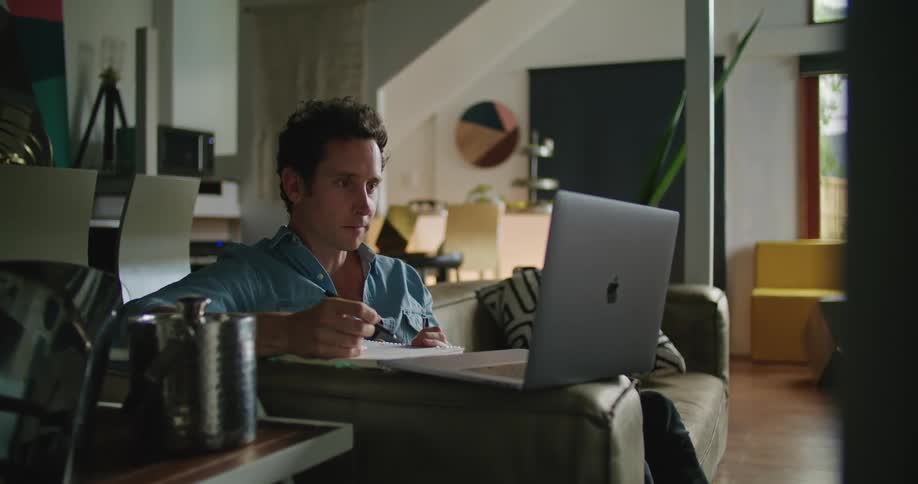 Man teleconferences via laptop laughing as he takes notes while sitting on couch in living room