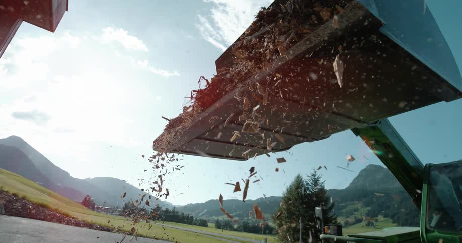 Wood chips fall from scoop arm of heavy machine parked on asphalt with mountains in the background