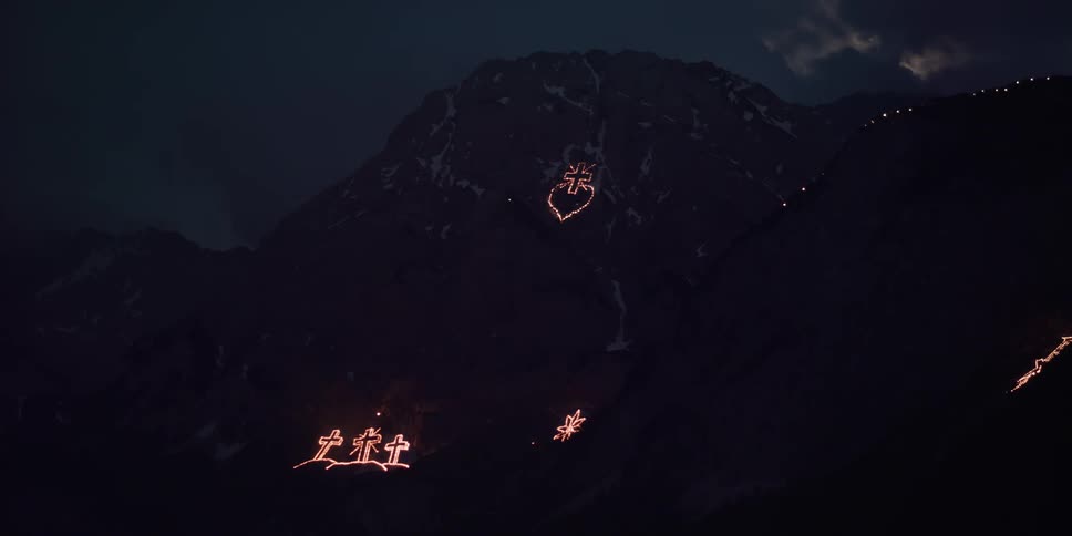 A lighting display of the cross of jesus appears with other light displays against a mountain at night