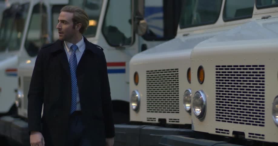 A man in an overcoat walks past a line of postal service trucks
