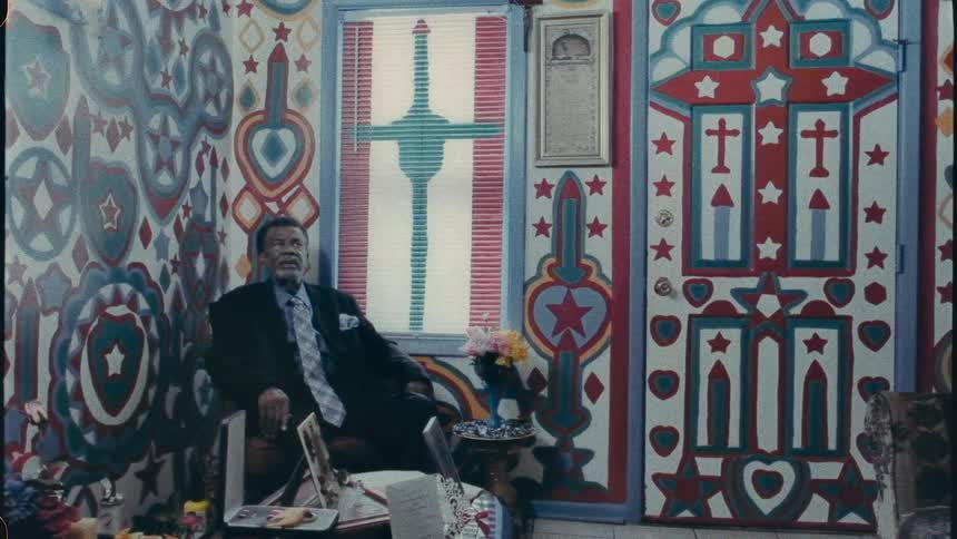 An old man is seated in a wall decorated room wearing a suit with a long tie