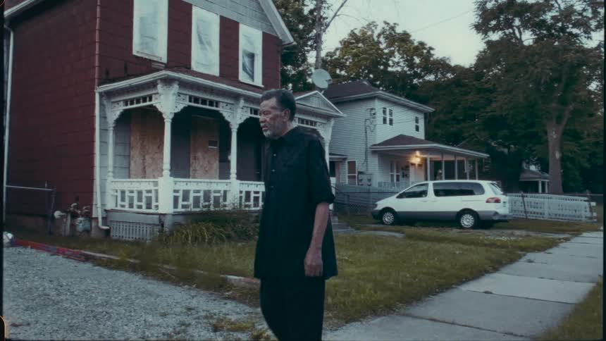 An old man in black walks down a suburban sidewalk toward a lavishly decorated artistic house and sculpture