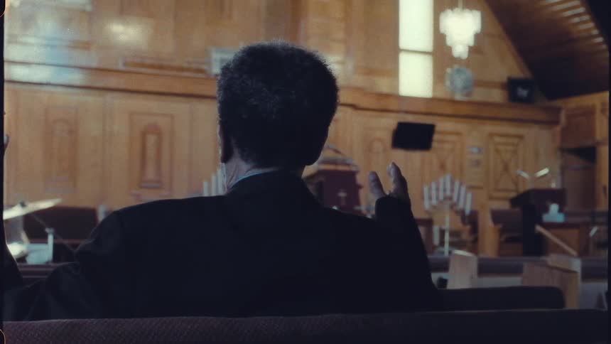 The backside of a man sitting in a church pew raising his hands up while facing forward