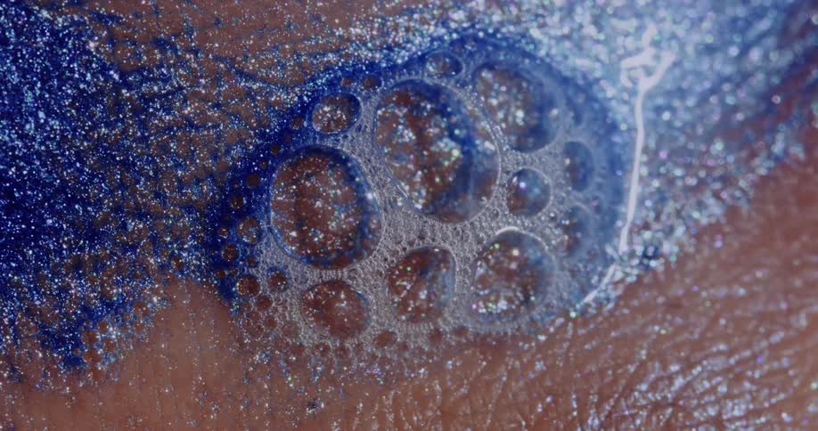 Light reflects on bubbles as a glittery blue liquid sparkles as it flows and twinkles on a pink surface