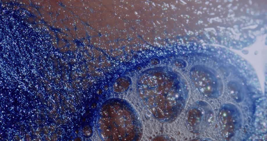 Light reflects on bubbles as a glittery blue liquid sparkles as it moves on a pink surface
