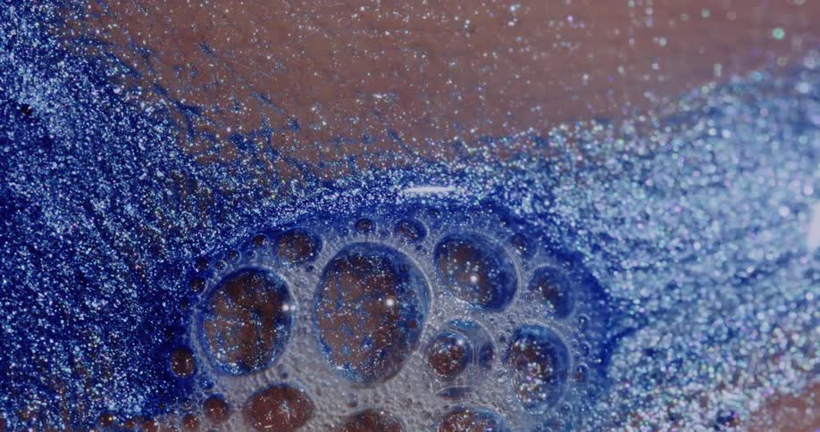 Light reflects on bubbles as a glittery blue liquid sparkles as it moves on a pink surface