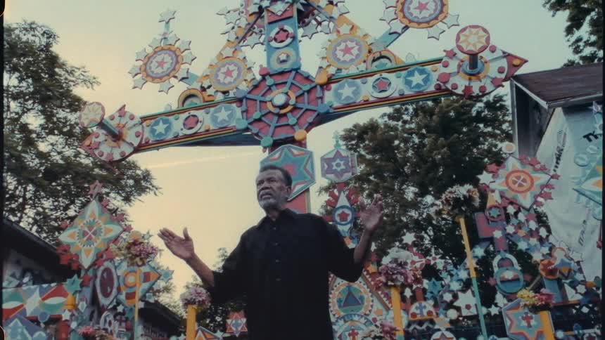 Man stands speaking with hands up and to his sides near colorful art displays including one in shape of cross