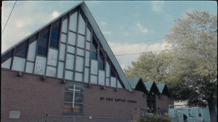 A cross is on the front of the building at mt erie baptist church as the trees move in the breeze near a house