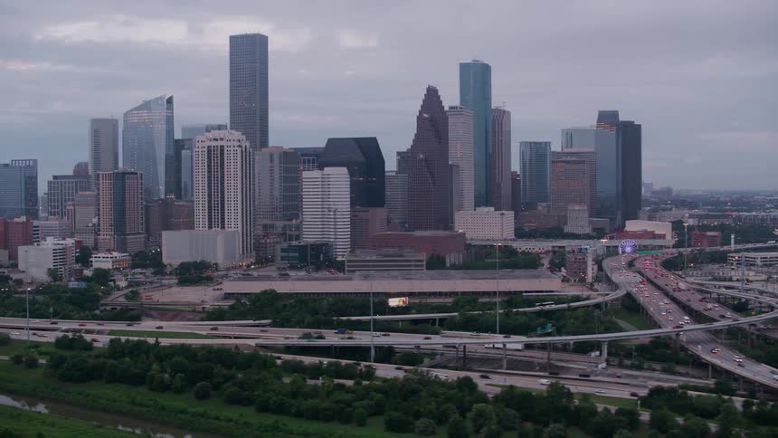 Traffic moves on a super highway junction around curves and bridges near a metropolitain city with tall buildings and skyscrapers under a cloudy sky