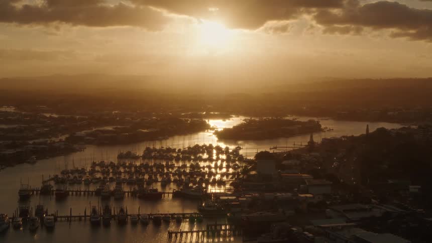 An aerial of boats docked at a coastal marina near a suburb neighborhood as the sun sets behind foggy clouds