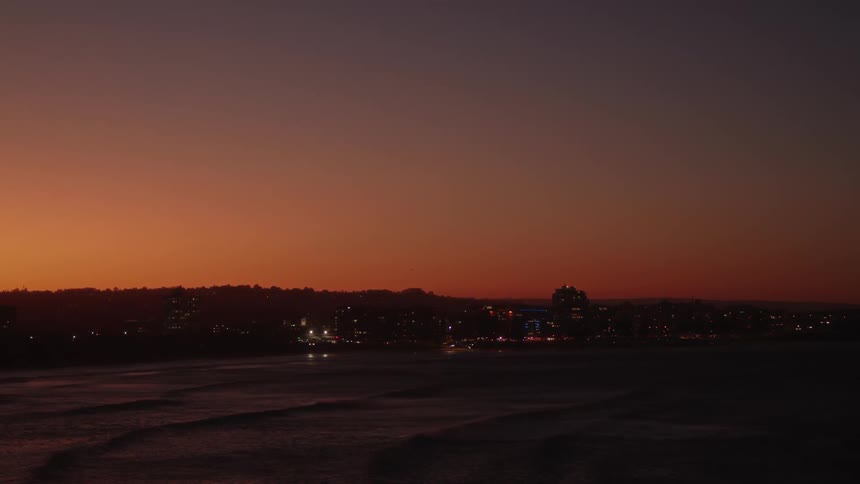 Waves roll into shore as lights on buildings along the coast glow under a red sunset sky