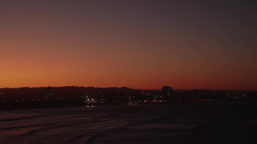 Drone view of a city along the coast lit up at dusk as the sun sets