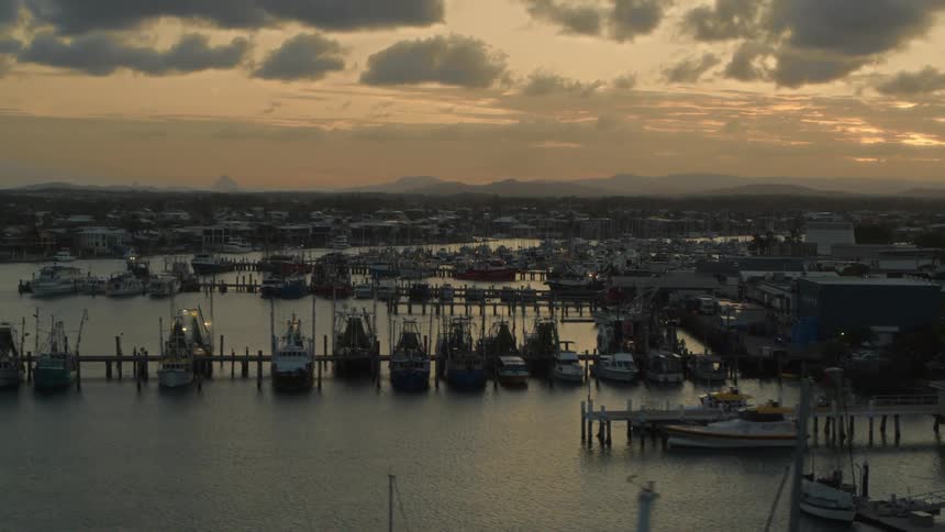 Town stretches from river with docked boats to misty mountains on horizon under a red tinted sky