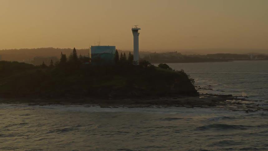 Building with beacon sits on promontory overlooking coast with city in the distance on foggy day