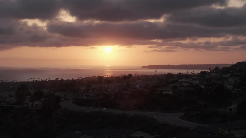The sun sets over the water in a colorful cloudy sky as traffic moves on neighborhood streets
