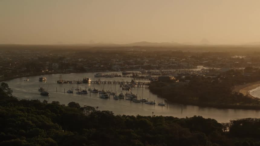 Boats are in a marina and near a pier in the lake off the coast of a town with a beach and trees along the shore with mountains in the distance under a hazy sky