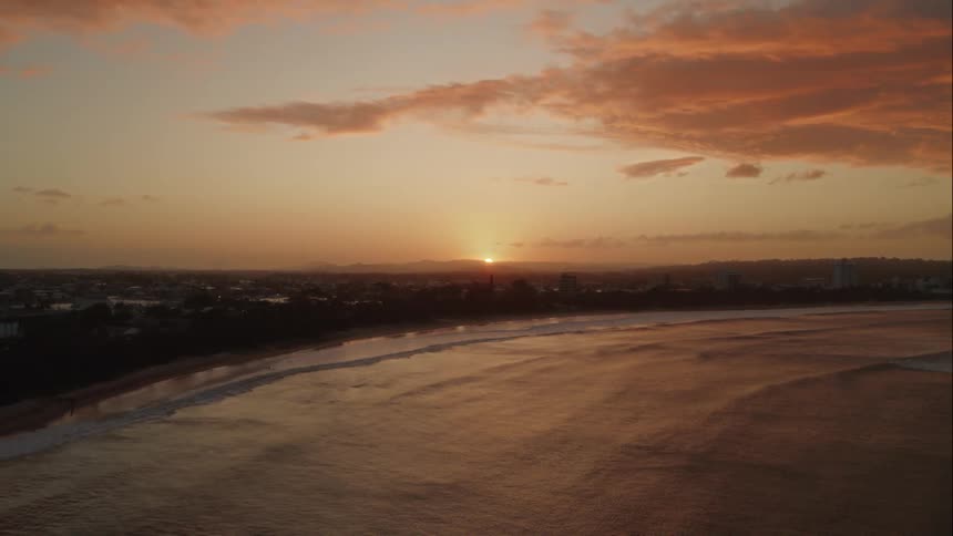 An aerial of waves rolling onto the beach with a suburban neighborhood behind the tree line as the sun sets on the horizon