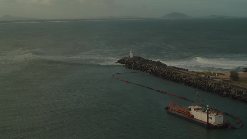 An aerial of a pier on the coast with a freight ship in the bay as waves crash against the pier rocks