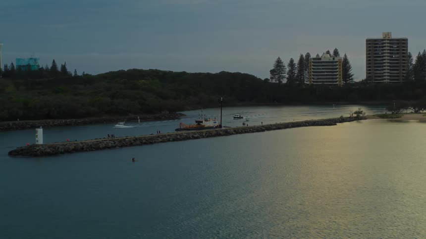 An aerial of a pier stretched onto the water as people walk on it at dusk while boats drive around in the harbor