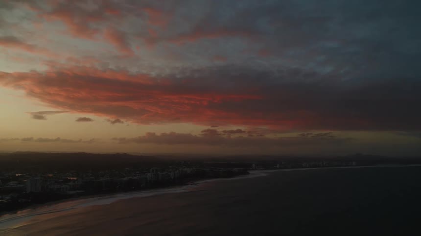 Waves roll into shore along the coast as colorful clouds float over buildings as the sun sets