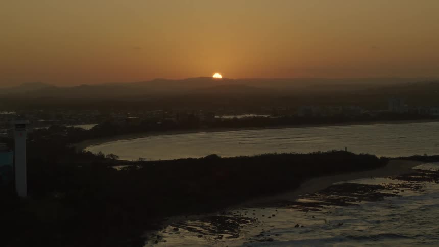 Sun sits on horizon a distance from coastal beacon on edge of city