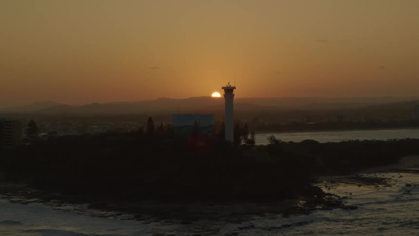 Sun sits on horizon under red sky in background a distance from beacon on edge of city on a foggy day