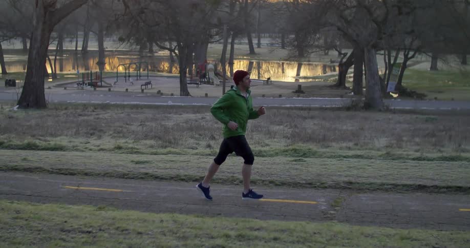 A man jogging down a track near a park with trees and a body of water