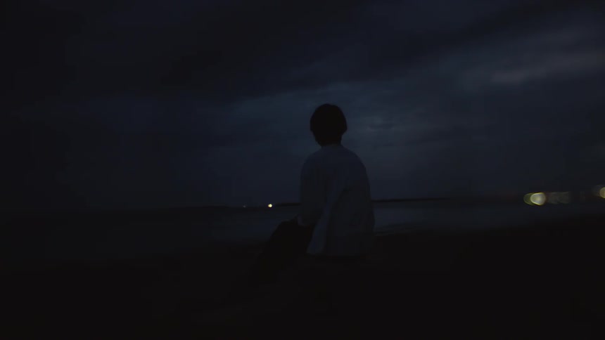 A person looking at the sea at night while lights shine in the distance