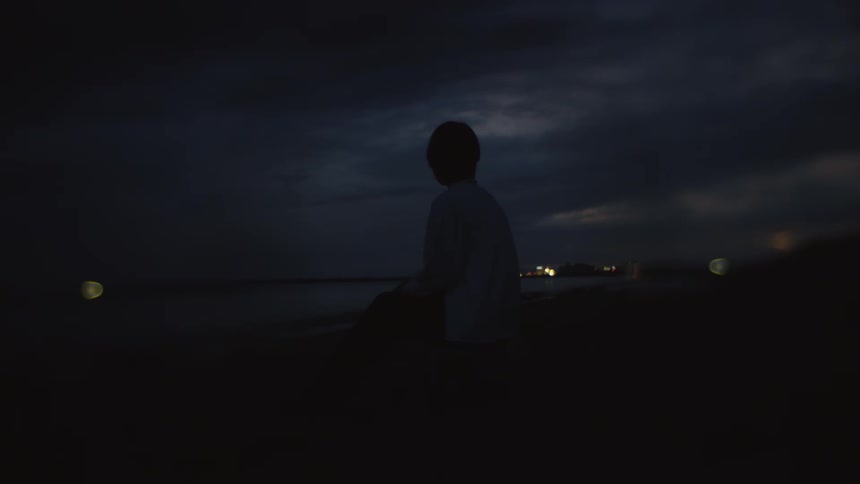 A person wearing a white shirt is standing near a rock along the coast facing the water in the darkness as lights glow in the distance