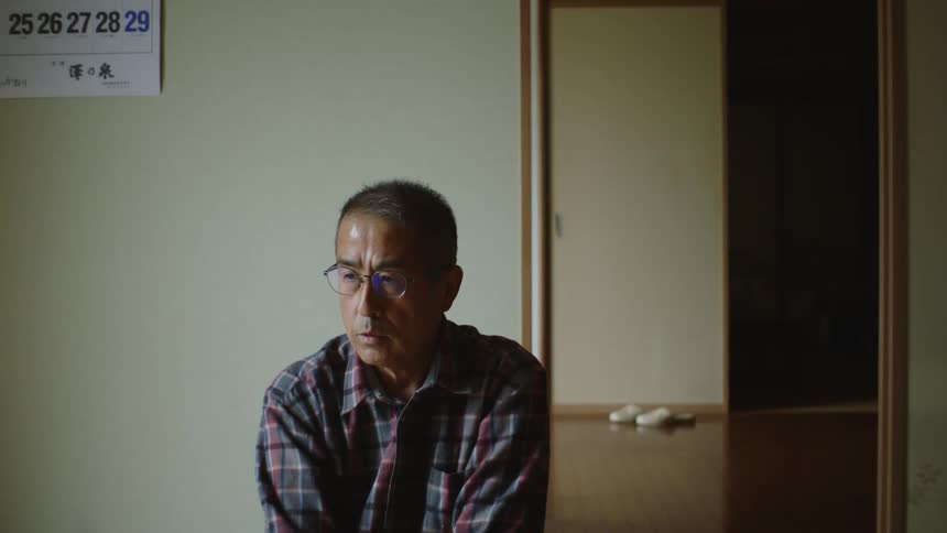 A man sitting in a room wearing glasses and a button up shirt while talking to someone