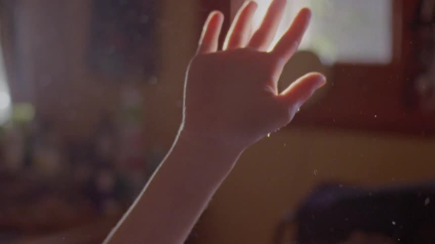 A persons hand raised up blocking light coming through a window displaying dust in the air