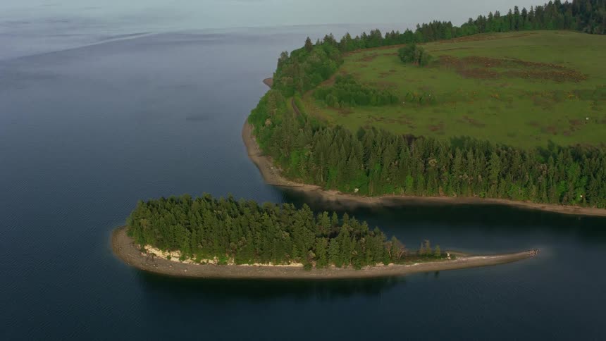 Trees cover an island with a beach around the edge and the peninsula has an open field surrounded by trees and woods