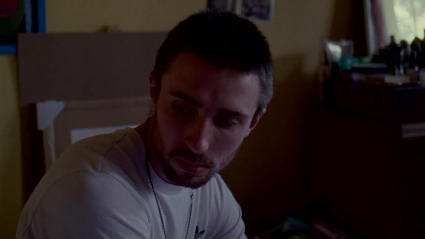 A man sitting with a sad expression on his face looking down toward the floor in a darker room