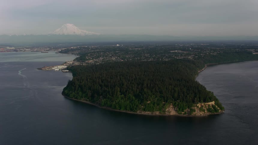 A tree covered peninsula as a ship sails in the water and there is a snow covered mountain in the far distance under a hazy sky