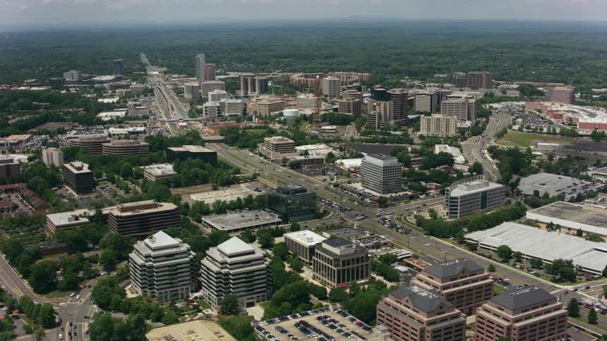 Traffic moves through a city with many buildings and cars in parking lots with trees and woods in the distance