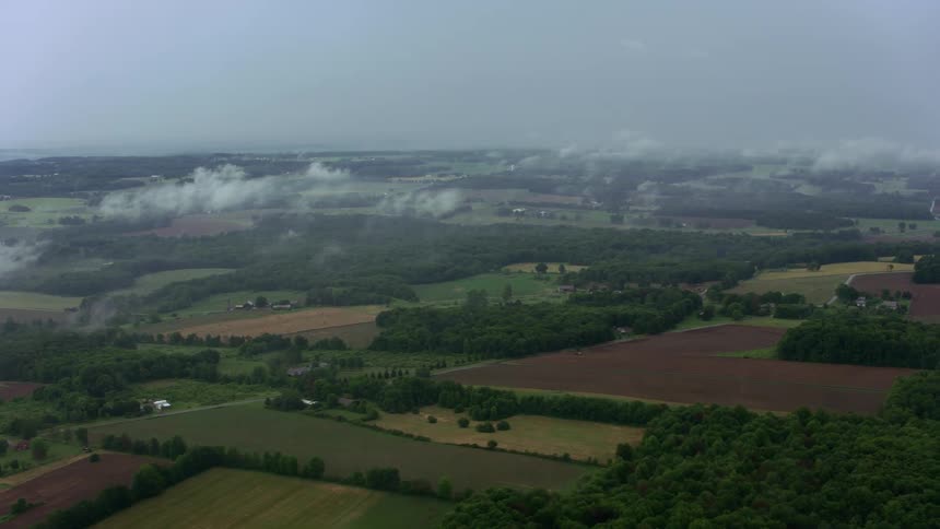Moving over a rural area with farms and houses spread far apart with areas of trees and woods as scattered clouds float in the sky