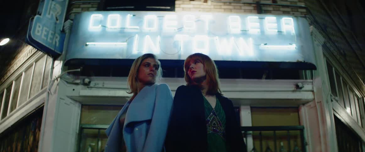 Two women wearing dresses and coats pose in front of a store at night