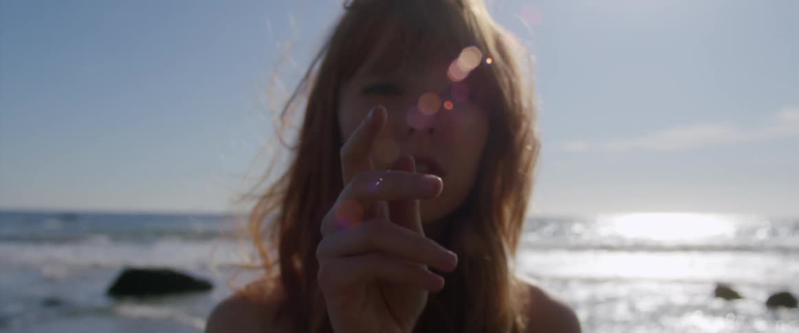 Woman on a beach reaches to the camera with her hand