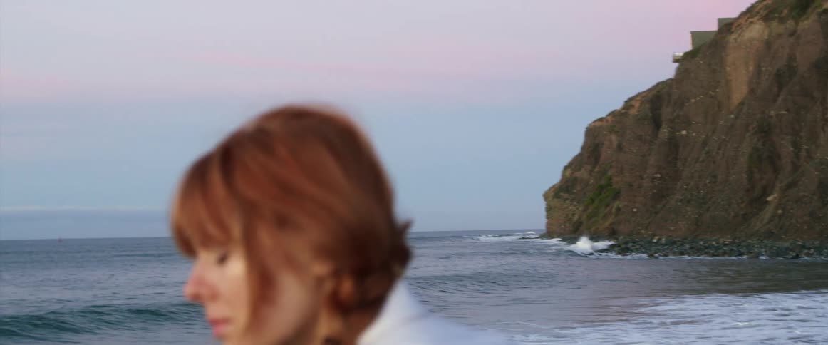 A young woman sitting at the beach stands up and looks off in to the distance