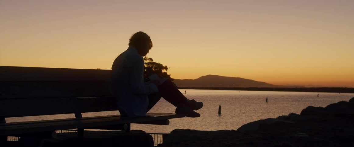Person sits on bench near water while horizon shows orange afterglow of setting sun