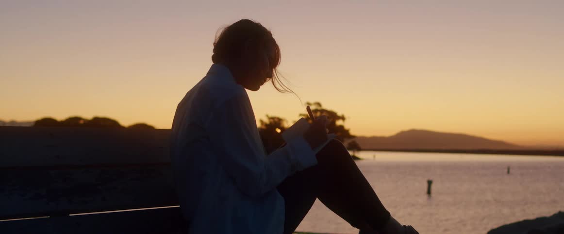 Silhouette of a young woman writing on a notebook next to the ocean at sunset