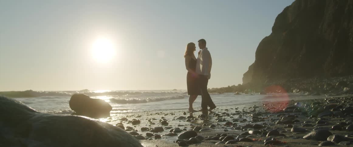 A man and woman in drenched clothing walk along a rocky beach while holding hands on a sunny day