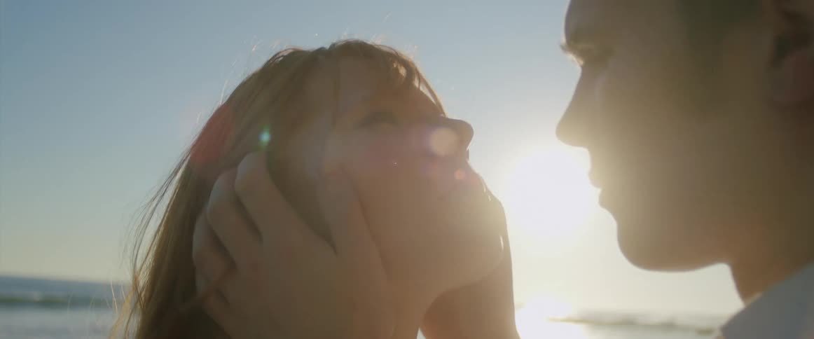 A woman looks up while the man leans in closer to her before kissing her as they stand on a beach