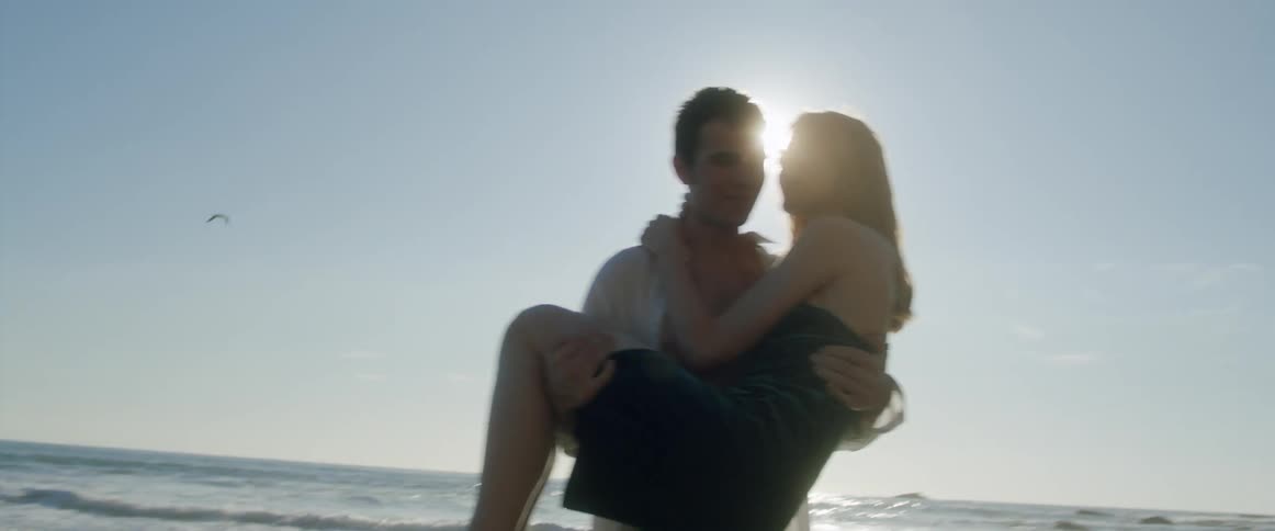 A man carries a woman from the ocean waters and she kisses him on the cheek