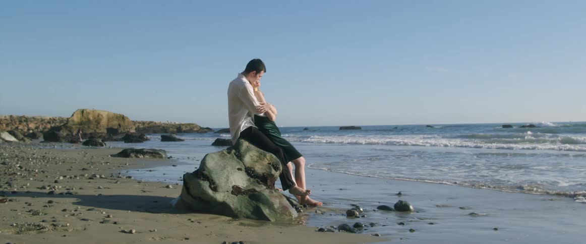 A man and woman sit together on a rock on the beach