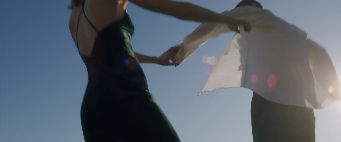 A man and woman hold hands while walking atop a row of tall rocks on a beach on a sunny day