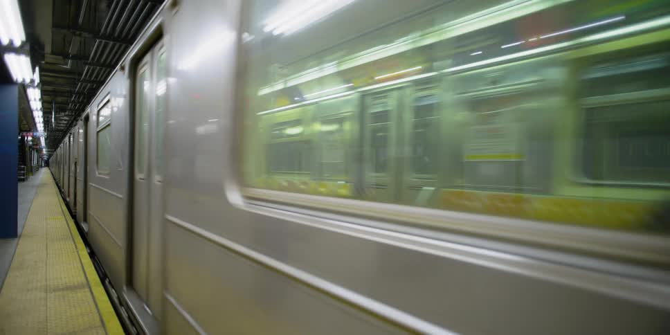 A single passenger sits on a subway train traveling past an empty platform in a station