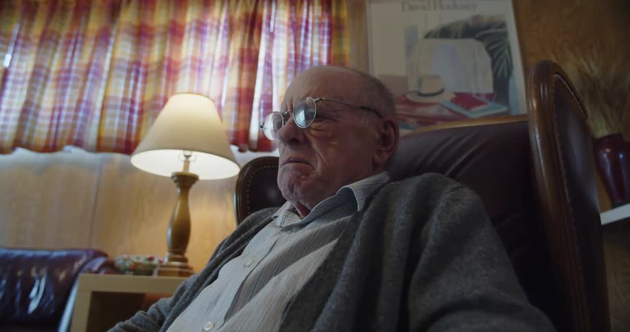 A man is sitting in a chair in his living room reading a newspaper